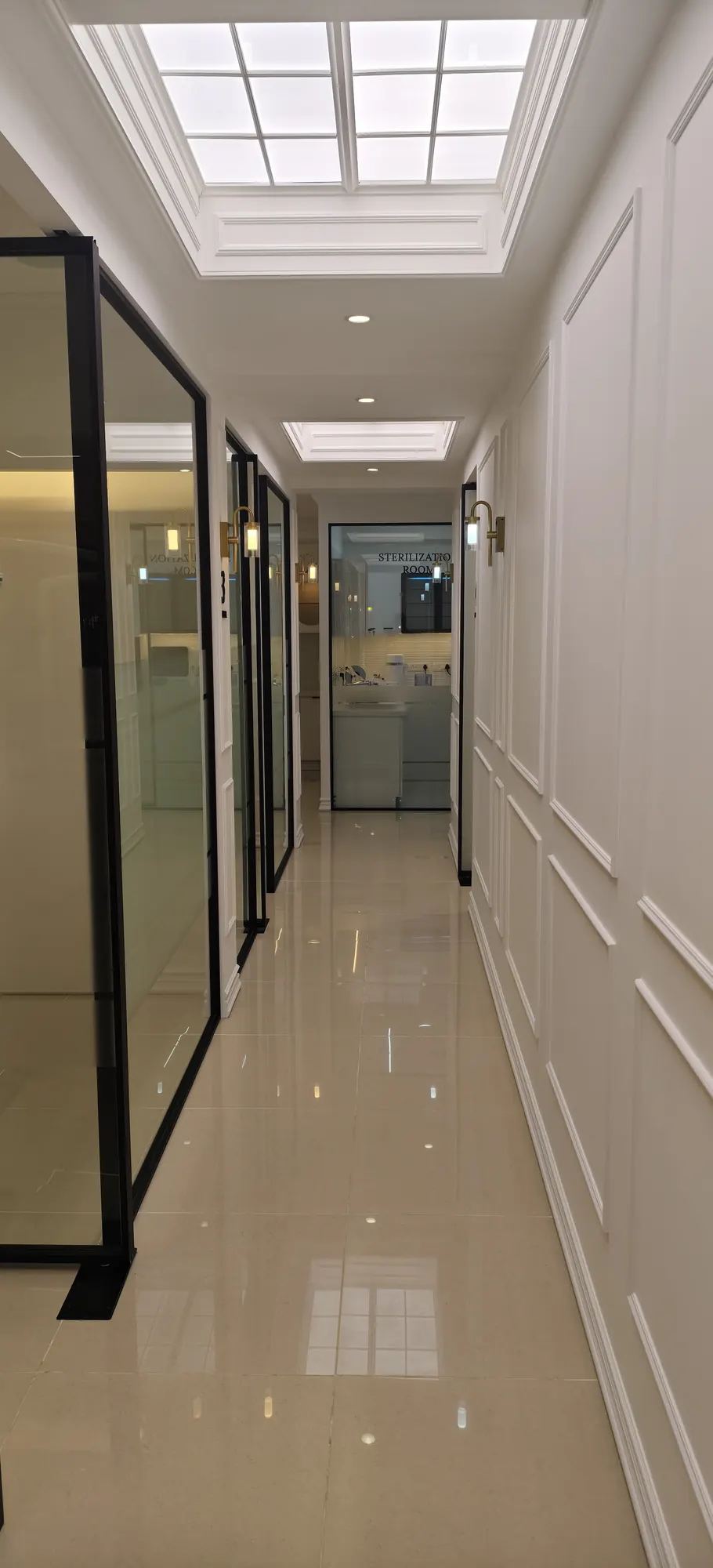A long white corridor under a skylight, lined with black-framed glass doors